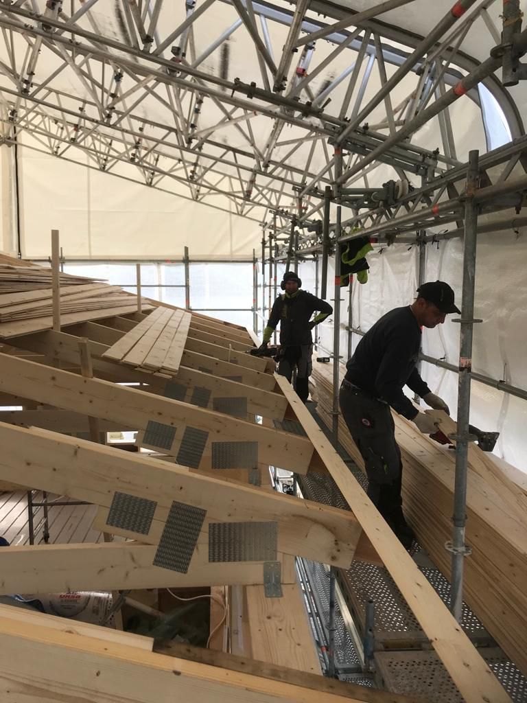 Två snickare i arbete vid takstolarna, stående på ställningen under väderskyddet.
